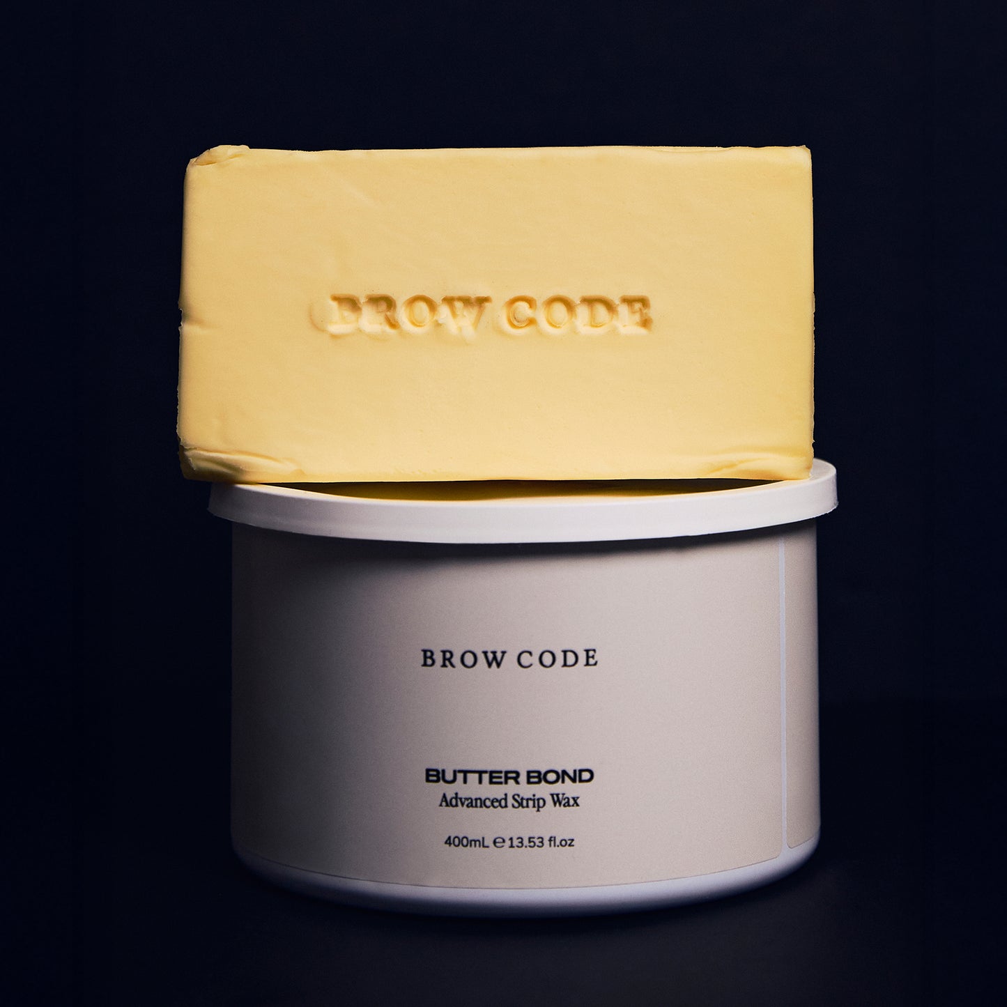 Close-up of a smooth, rectangular block of Butter Bond wax in its solid form, set against a dark navy background. The creamy yellow tone and clean edges highlight its buttery, hypoallergenic formula